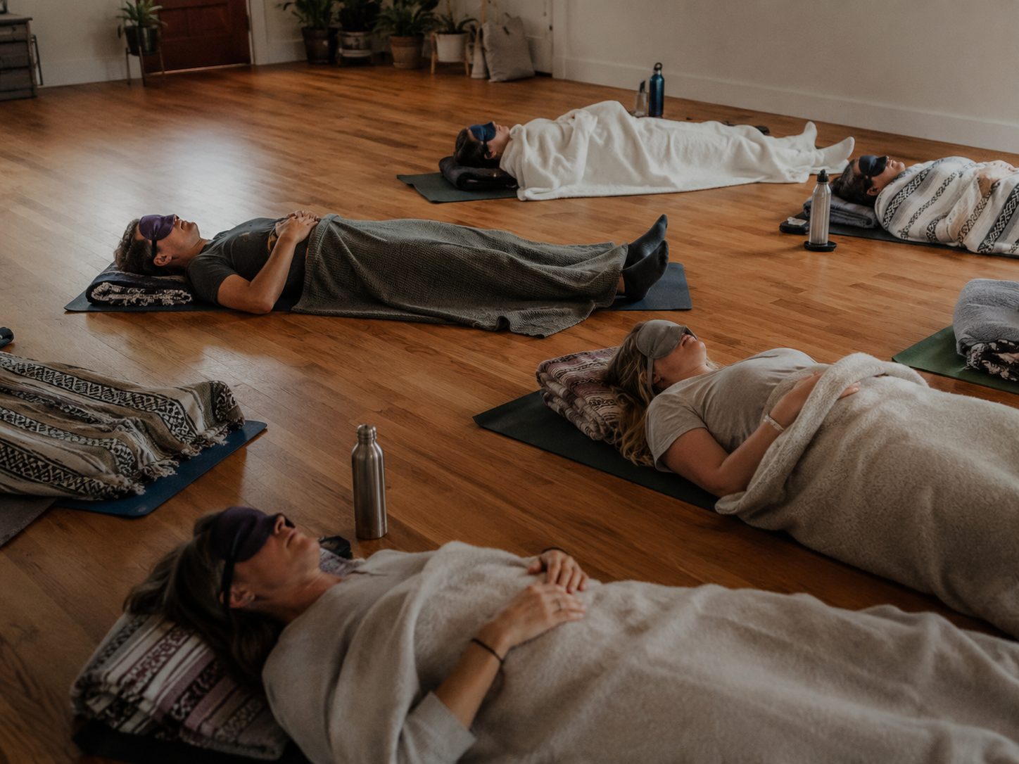Participants rest on mats under blankets with eye masks, in a bright indoor space with a wooden floor.